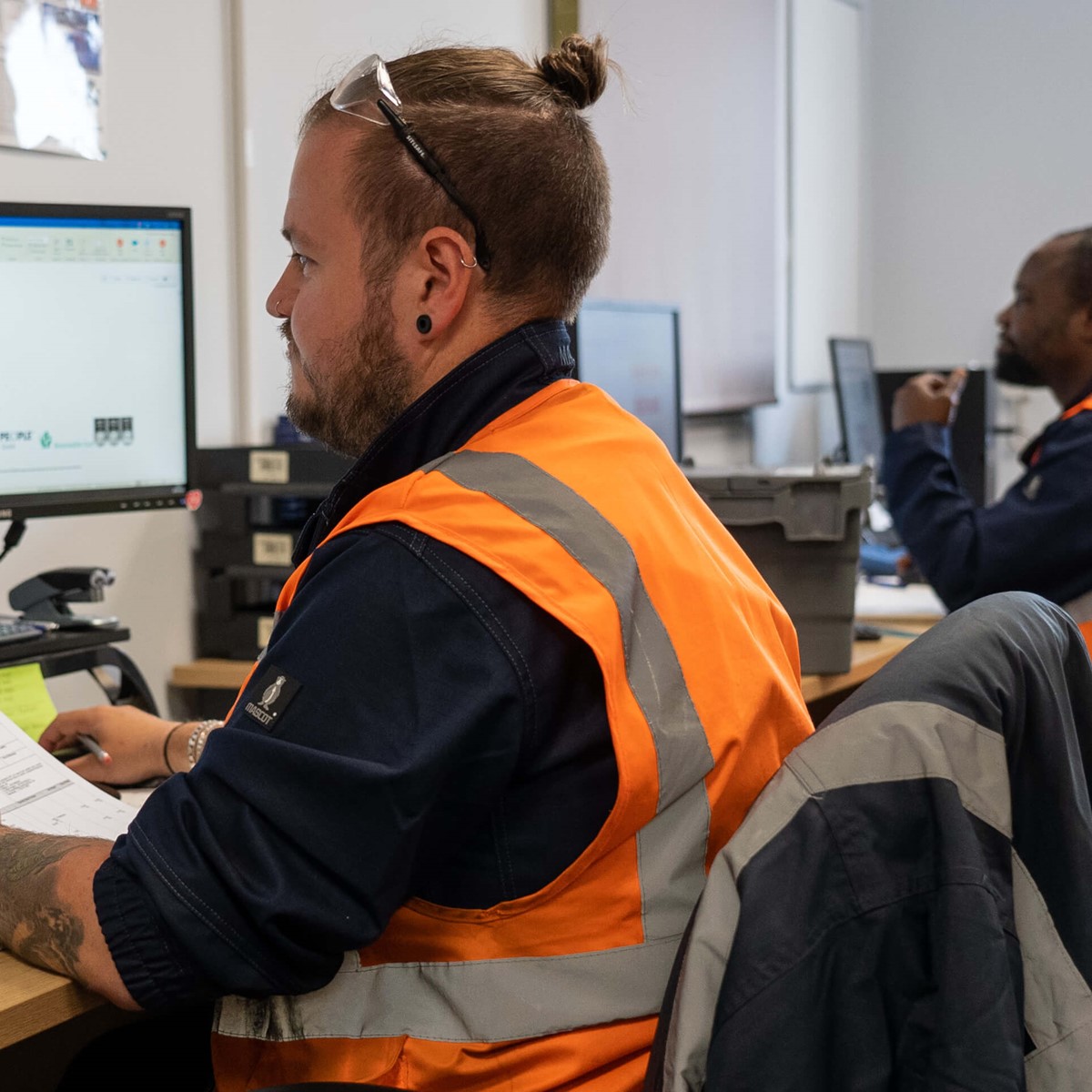 Man in high-vis jacket holds paper while working at a computer